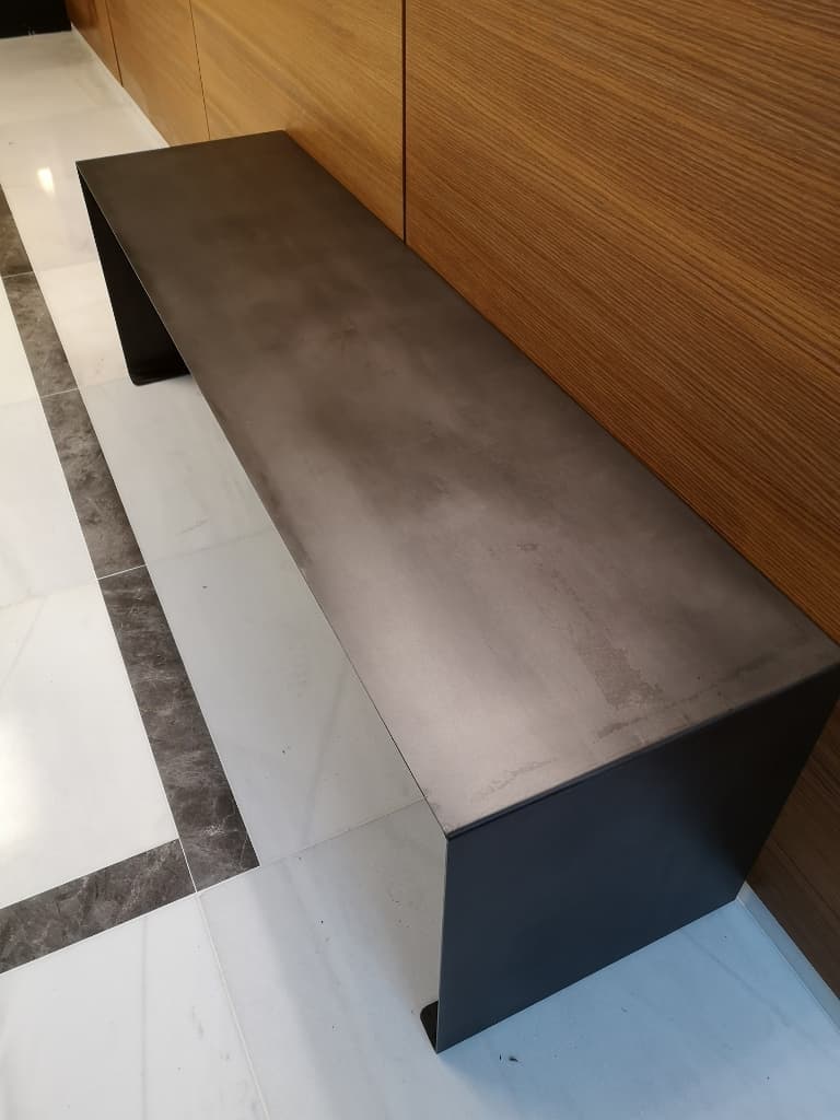 Banc minimaliste en métal patiné anthracite adossé à un mur en boiserie dans un hall d'entrée moderne.
