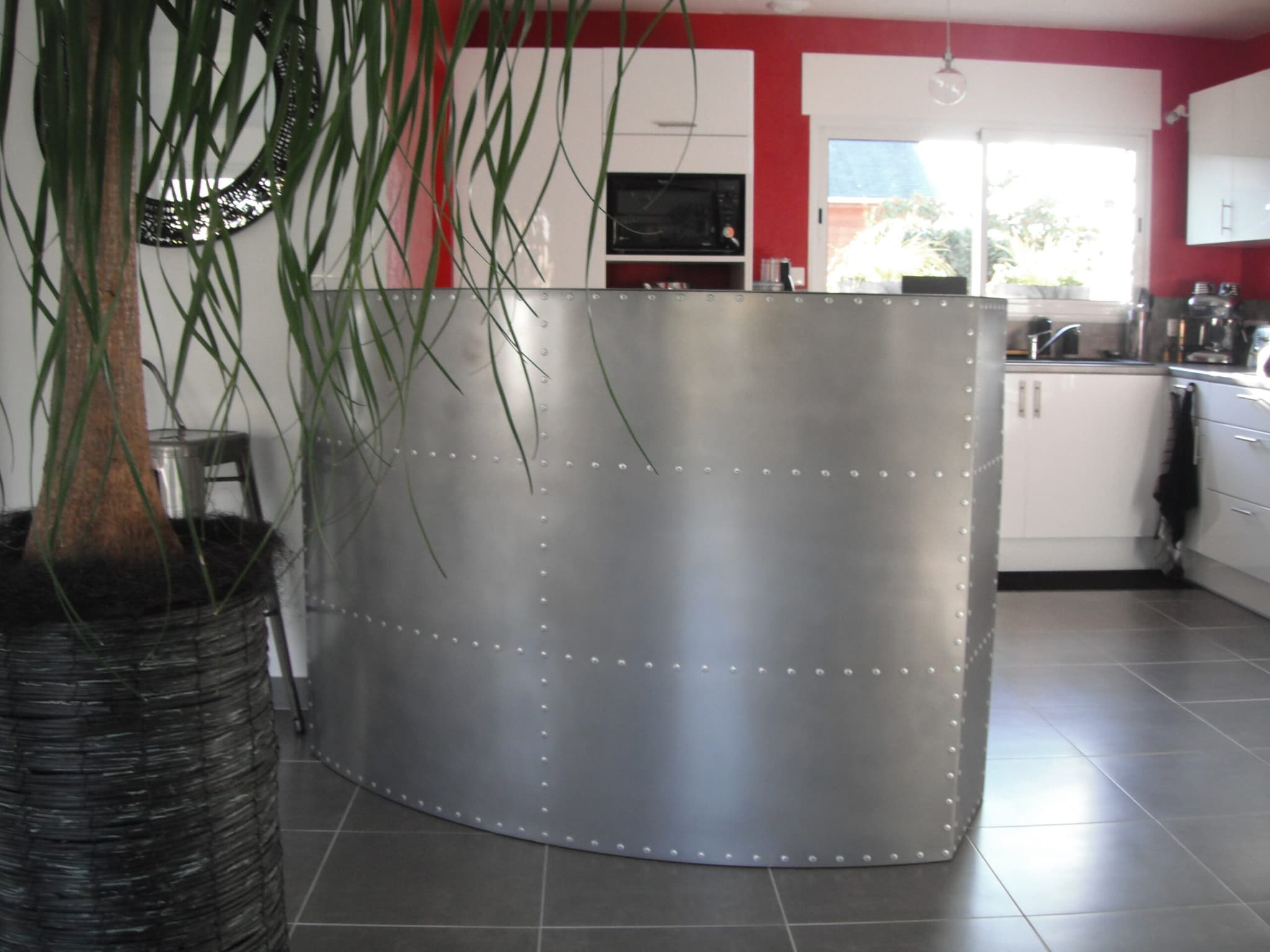 Grand comptoir de bar sur mesure en métal riveté au design courbé, séparant une cuisine moderne d'une pièce à vivre.
