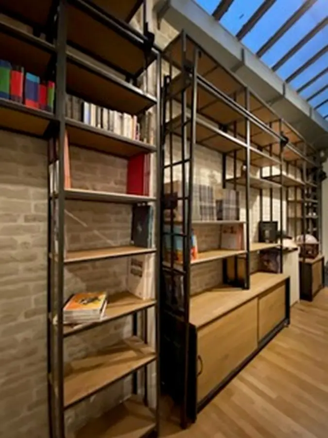 Étagères industrielles en métal et bois longent un mur en pierre, regorgeant de livres et objets, avec un meuble de rangement en bois au sol.