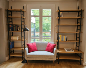 Bibliothèque en bois et métal noir autour d’un canapé gris avec coussins rouges près d’une grande fenêtre.