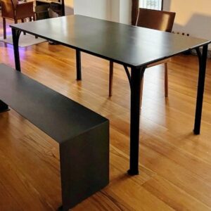 Table à manger Baltard en acier brut calaminé noir avec piètement arqué et banc assorti sur un parquet en bois.