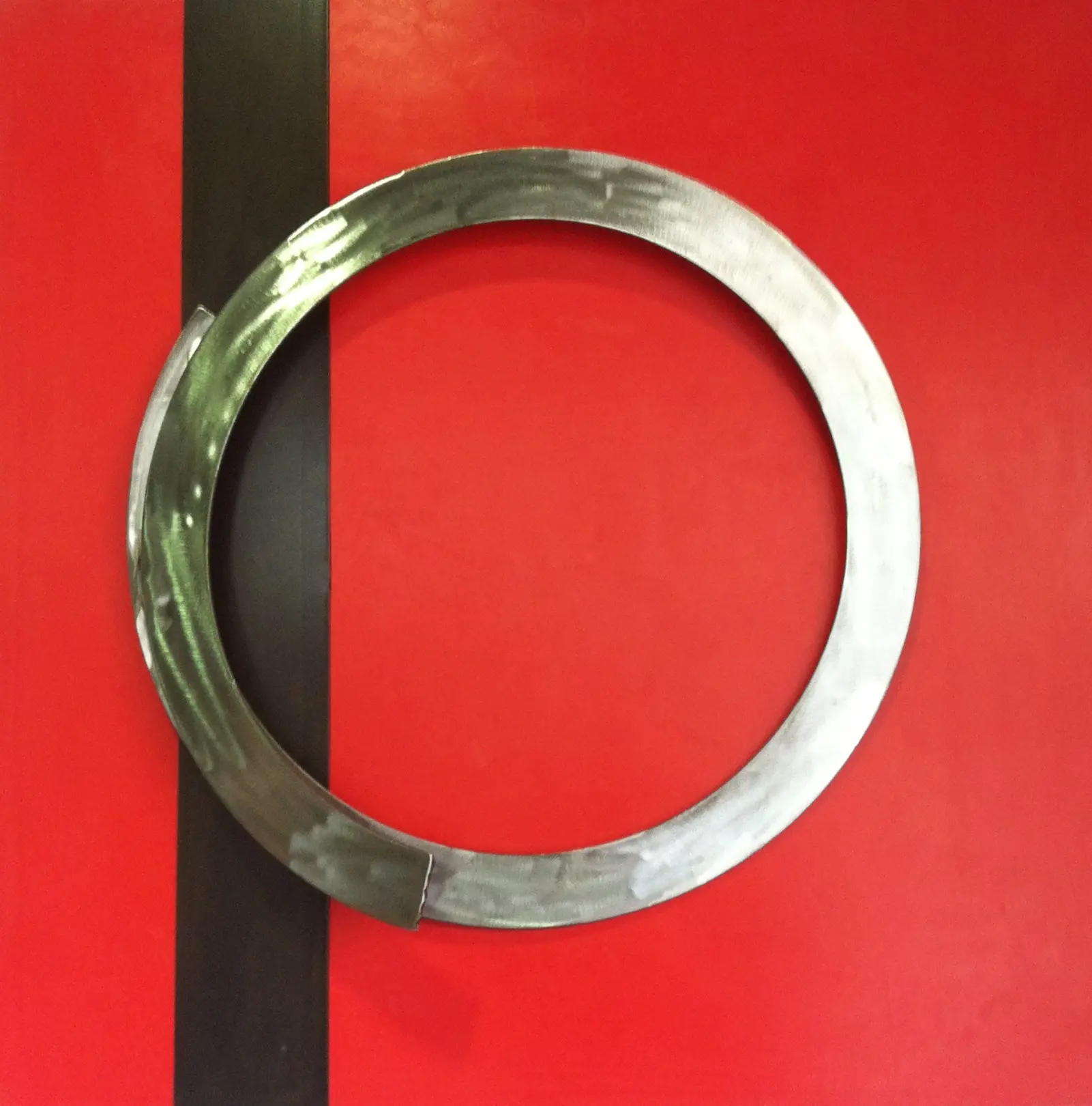 Anneau métallique décoratif à patine brute posé sur un fond rouge, avec une bande verticale noire en arrière-plan.